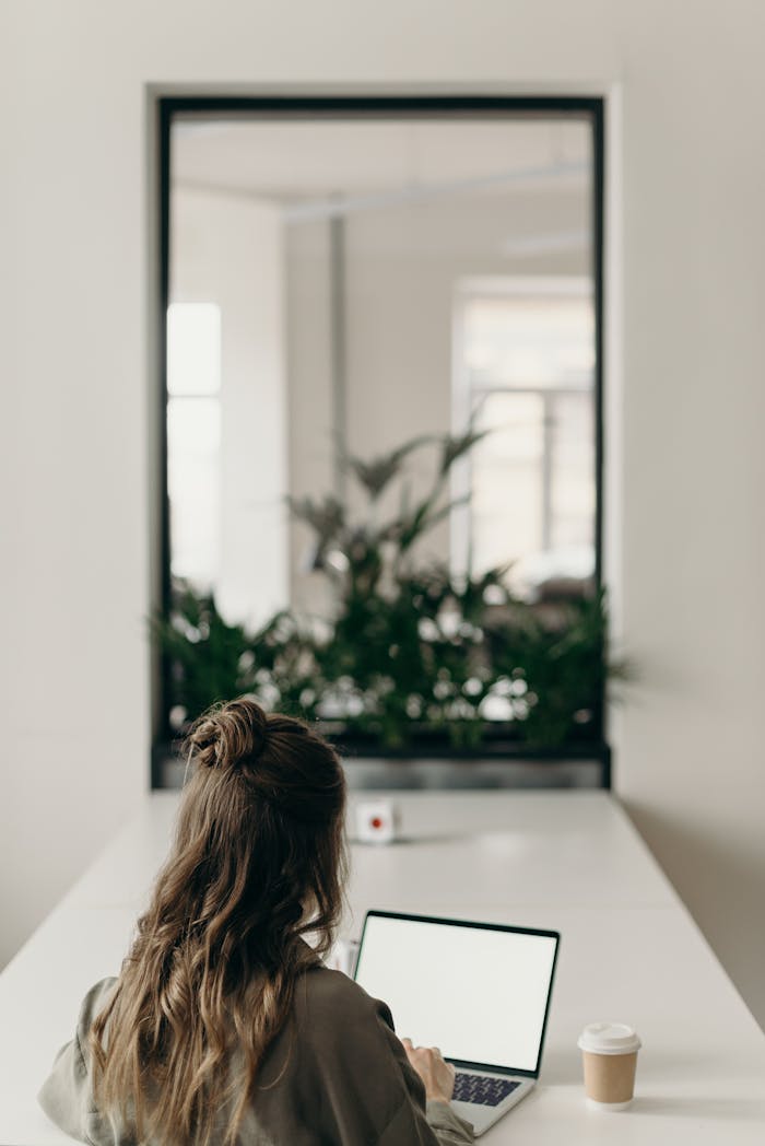 story-01 A woman with curly hair works on a laptop, facing a window with plants, symbolizing remote work.